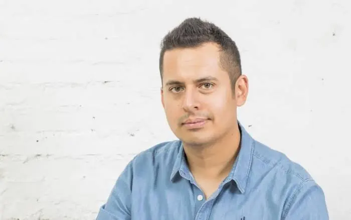 Retrato del fundador y creador de la herramienta SEO DinoRank, un hombre con camisa de mezclilla azul, junto a una placa conmemorativa en una pared de ladrillo blanco.
